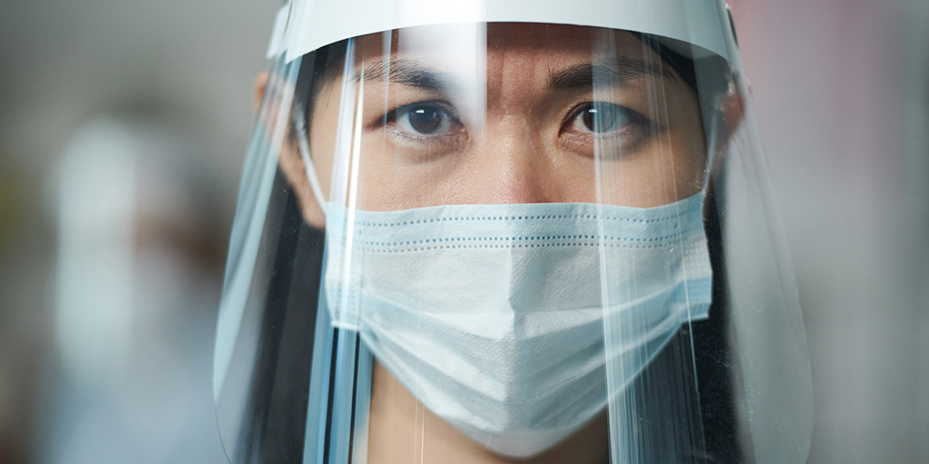Young serious nurse in uniform and protective workwear looking at you through transparent mask